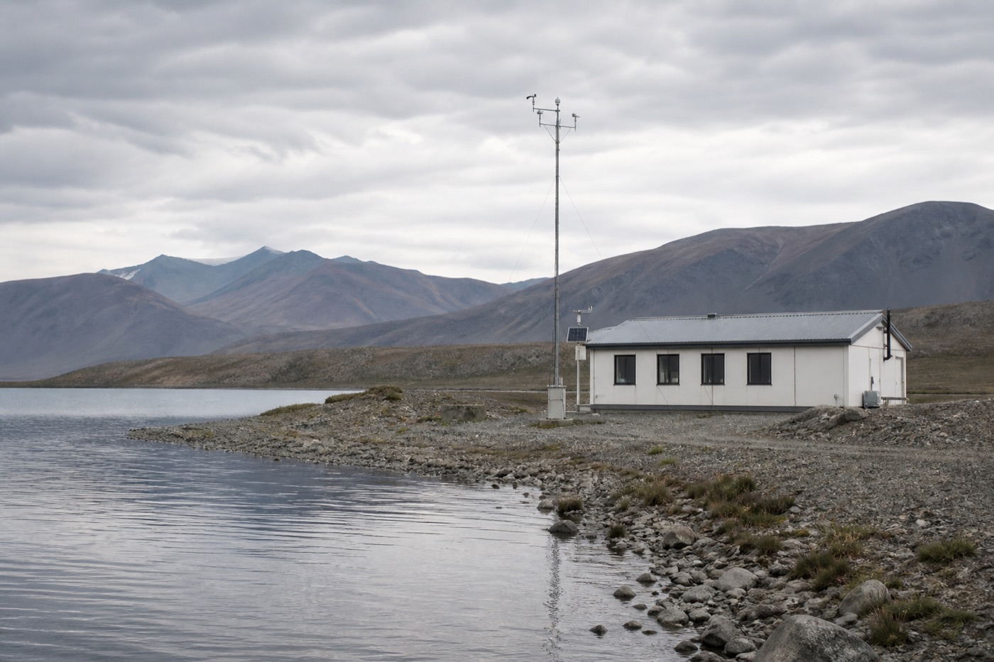 栖澜湖南岸砾石湖滩上的单层观测站建筑,一旁立有装载小型仪器的气象观测桅杆,远处是灰绿色山脉